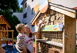 Mutter trägt den Sohn auf dem Arm und zeigt auf die Insekten-Jugendherberge.