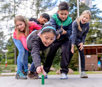 Eine Gruppe von fünf Kindern führt eine Kooperationsübung im Freien durch, bei der sie eine Mitschülerin gemeinsam halten, damit sie einen Baustein am Boden erreicht.